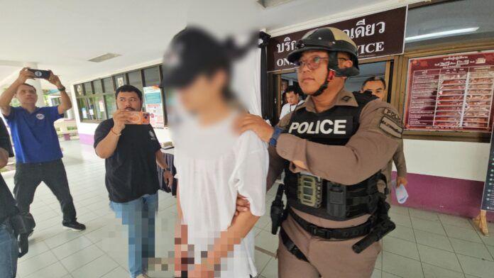 Pattaya news photo of a Thai police officer in a POLICE vest and helmet escorting the blurred Chinese suspect through a police station in Chonburi after the Huai Yai weapons cache was seized